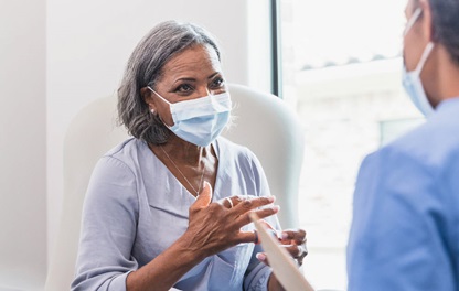 A photograph of an older woman wearing a surgical facemask gesturing while talking to a health care provider