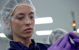 Image of a chemist wearing a hairnet, protective eye protection, and latex gloves, examining a small syringe of liquid