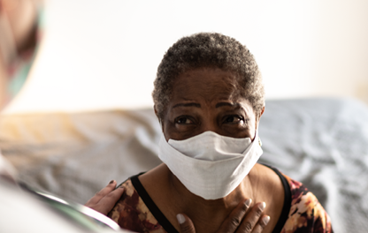 A photograph of an older African American woman wearing a facemask, seated, looking at a doctor standing in the foreground