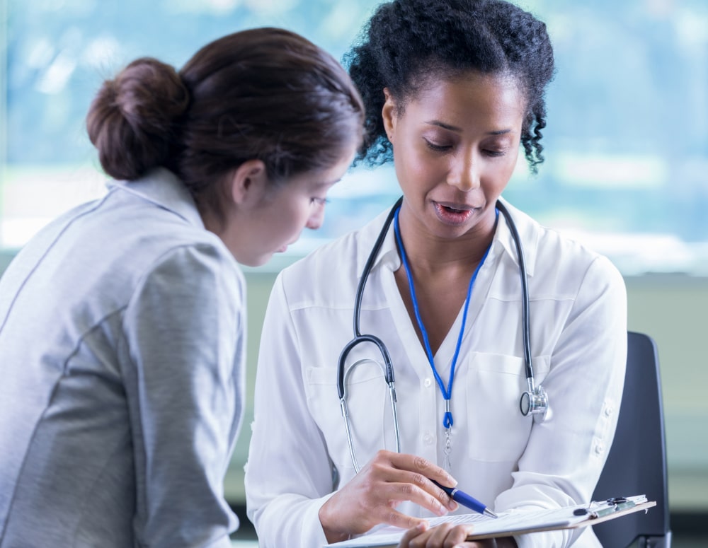 Female doctor wearing a stethoscope and pointing at a clipboard with a pen, talking with a female patient