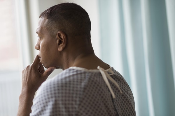 African American male patient wearing a hospital gown looking out a window, with his hand poised over his chin in a contemplative, concerned expression