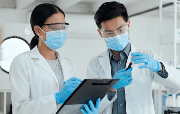 Scientists in lab coats with masks on working in lab looking at vial.