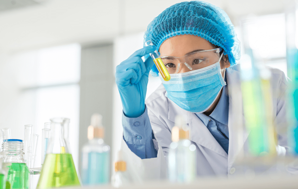 Pharmaceutical lab tech wearing gloves, hair covering, glasses, and a face mask, intently examining a test tube of yellow liquid