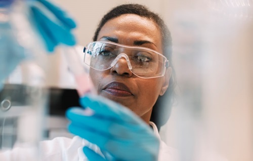 Close-up photograph of an African-American female scientist's blue-gloved hands handling a test tube, with her intently focused expression in focus